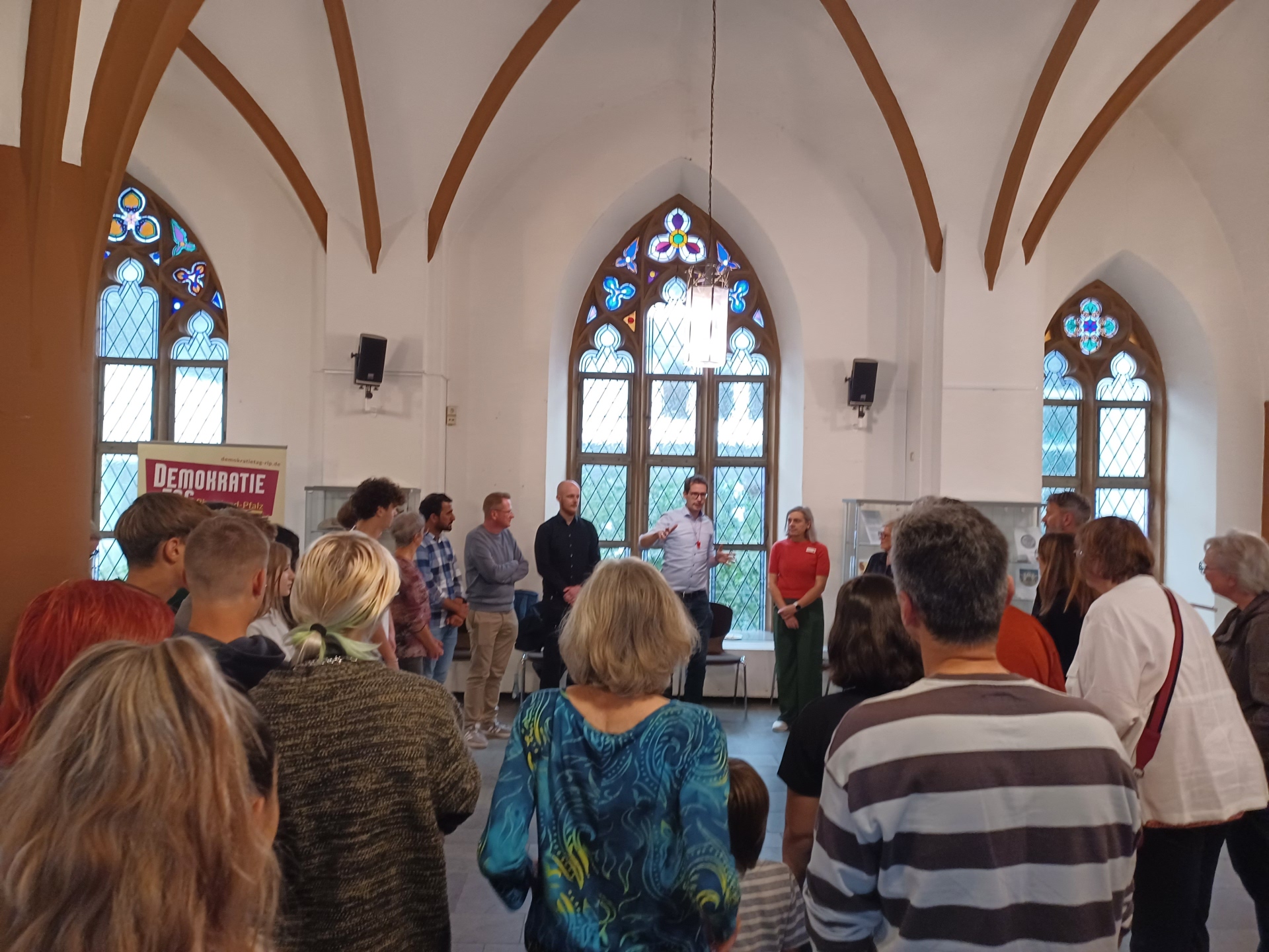 Eine Gruppe von Menschen steht in einem Raum mit hohen, gotischen Fenstern und hört einem Mann zu, der vorne spricht und gestikuliert. Neben ihm stehen mehrere Personen, darunter eine Frau in rotem Oberteil, und im Hintergrund ist ein Roll-Up mit der Aufschrift „Demokratie leben!“ sichtbar.