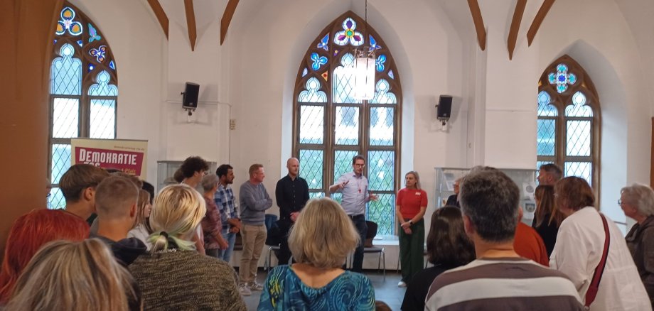 Eine Gruppe von Menschen steht in einem Raum mit hohen, gotischen Fenstern und hört einem Mann zu, der vorne spricht und gestikuliert. Neben ihm stehen mehrere Personen, darunter eine Frau in rotem Oberteil, und im Hintergrund ist ein Roll-Up mit der Aufschrift „Demokratie leben!“ sichtbar.