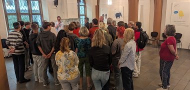 Eine Gruppe von etwa 30 Menschen steht dicht gedrängt in einem Raum mit hohen Fenstern und gotischen Bögen, während ein Mann vorne spricht und offenbar eine Führung oder Präsentation hält. Die Atmosphäre wirkt aufmerksam und interessiert, im Hintergrund hängen Plakate mit grafischen Illustrationen und deutschen Begriffen wie „Meinungsfreiheit“.
