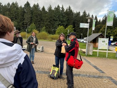 Mehrere Personen stehen auf einem gepflasterten Platz am Waldrand, eine Frau überreicht einem Mann mit roter Umhängetasche eine Papiertüte. Im Hintergrund stehen Wegweiser, Infotafeln und grüne Waldlandschaft.