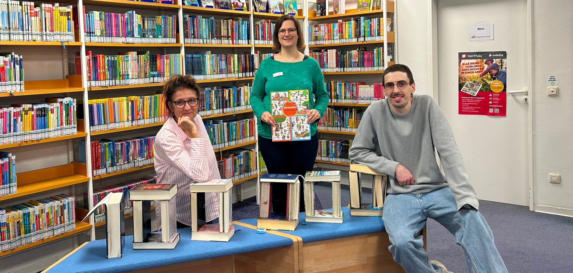 Drei Personen posieren in einer farbenfrohen Kinder- und Jugendbuchabteilung einer Bibliothek. Auf der blauen Sitzbank vor ihnen steht mit aufgeschlagenen Büchern das Wort „1000“ aufgebaut. Die Frau in der Mitte hält ein großformatiges Wimmelbuch mit dem Titel „Willkommen in Wimmelstadt“ in den Händen.