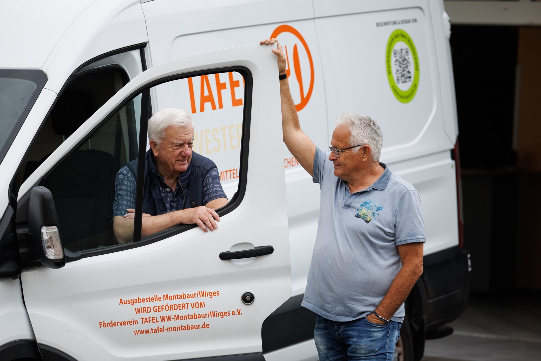 Zwei ältere Männer unterhalten sich freundlich an einem weißen Lieferwagen der Tafel Montabaur/Wirges. Einer sitzt am Steuer mit offener Tür, der andere lehnt sich entspannt mit einem Arm an die Tür, während auf dem Wagen das orangefarbene Tafel-Logo und Förderinformationen zu lesen sind.