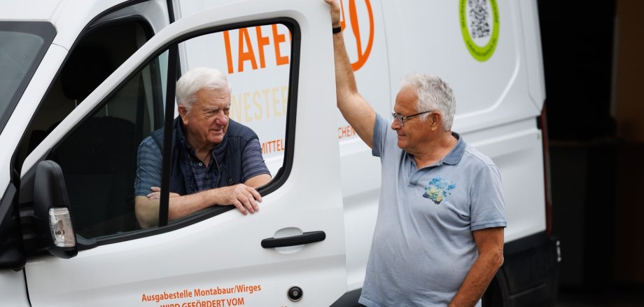 Zwei ältere Männer unterhalten sich freundlich an einem weißen Lieferwagen der Tafel Montabaur/Wirges. Einer sitzt am Steuer mit offener Tür, der andere lehnt sich entspannt mit einem Arm an die Tür, während auf dem Wagen das orangefarbene Tafel-Logo und Förderinformationen zu lesen sind.