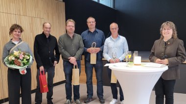 echs Personen stehen bei einer Feier in der Halle zusammen; eine Frau hält einen Blumenstrauß, eine andere ein Mikrofon.