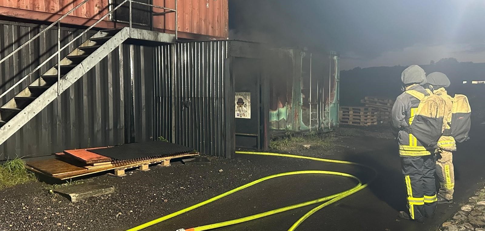 Zwei Feuerwehrangehörige in Schutzkleidung und mit Atemschutzgeräten stehen vor einer Übungsanlage. Feuerwehrschläuche liegen auf dem Boden.