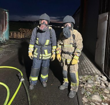 Zwei Feuerwehrangehörige stehen in Schutzkleidung und mit angelegten Atemschutzmasken vor der Übungsanlage.