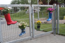 Das Bild zeigt ein metallenes Eingangstor, dekoriert mit Blumen in Gummistiefeln